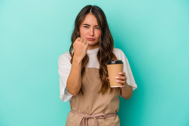 Young caucasian store clerk woman holding a takeaway coffee isolated on blue background showing fist to camera, aggressive facial expression.