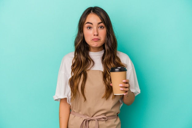 青い背景に分離されたテイクアウトコーヒーを保持している若い白人の店員の女性は肩をすくめ、目を開けて混乱します。