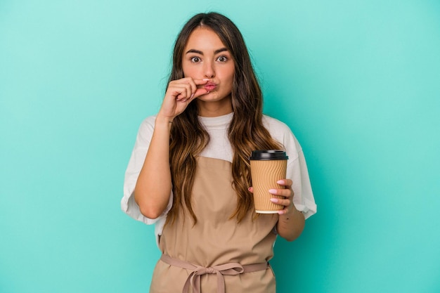 Young caucasian store clerk woman holding a takeaway coffee isolated on blue background with fingers on lips keeping a secret.