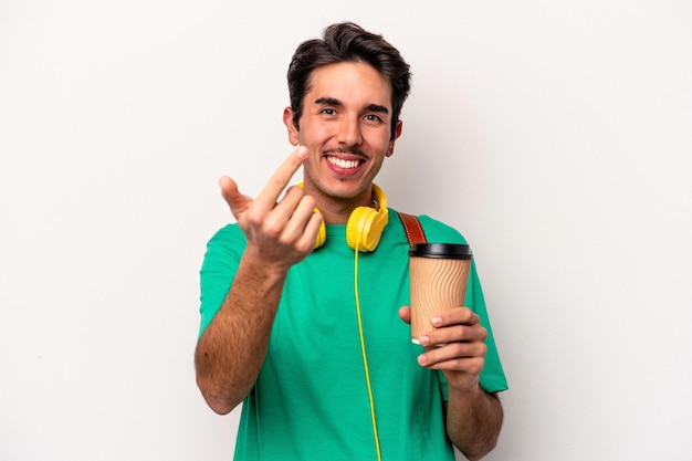 Young caucasian student man drinking coffee isolated on white background pointing with finger at you as if inviting come closer.