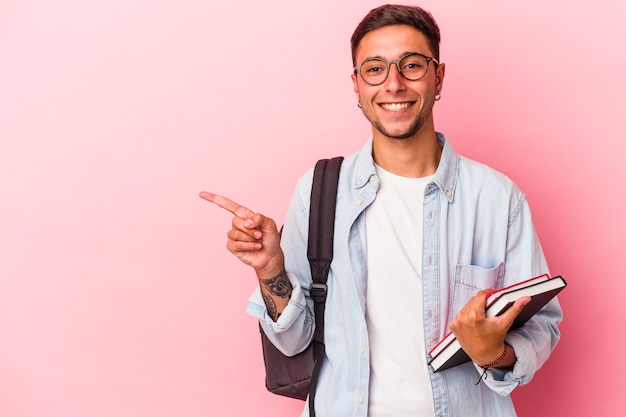 ピンクの背景に分離された本を持っている若い白人学生の男性は、笑顔で脇を指して、空白のスペースで何かを示しています。