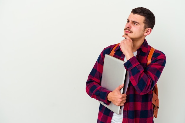 Uomo giovane studente caucasico che tiene un computer portatile isolato sul muro bianco che guarda lateralmente con espressione dubbiosa e scettica.