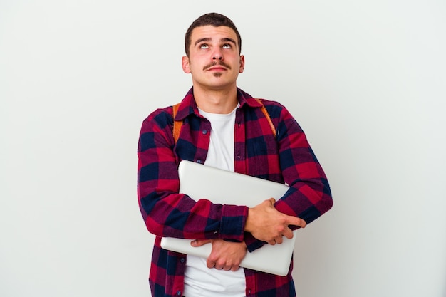 Young caucasian student man holding a laptop isolated on white wall tired of a repetitive task.