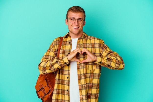 Young caucasian student man isolated on blue smiling and showing a heart shape with hands.