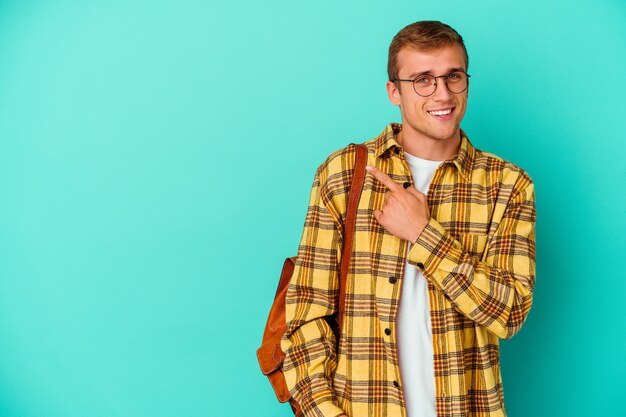 Young caucasian student man isolated on blue wall smiling and pointing aside, showing something at blank space.