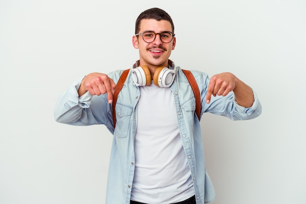 白い背景で隔離の音楽を聴いている若い白人学生の男は、指で下向き、前向きな気持ち。