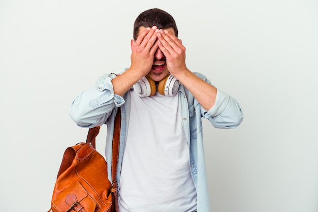 Young caucasian student man listening to music isolated on white wall covers eyes with hands, smiles broadly waiting for a surprise