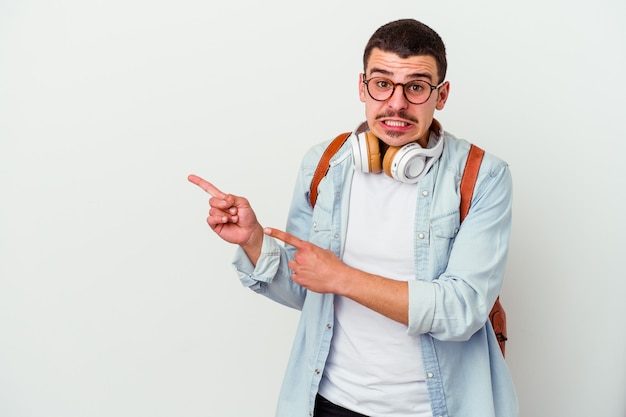 Young caucasian student man listening to music isolated on white wall shocked pointing with index fingers to a copy space.