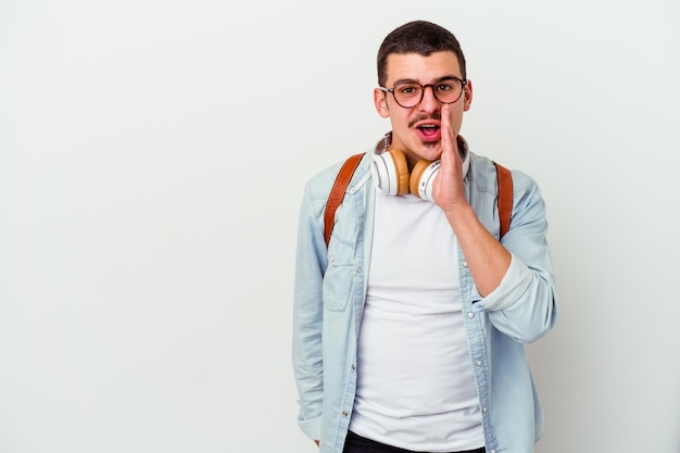 Young caucasian student man listening to music on white is saying a secret hot braking news and looking aside