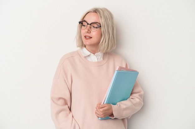 Young caucasian student woman holding books isolated on white background looks aside smiling, cheerful and pleasant.