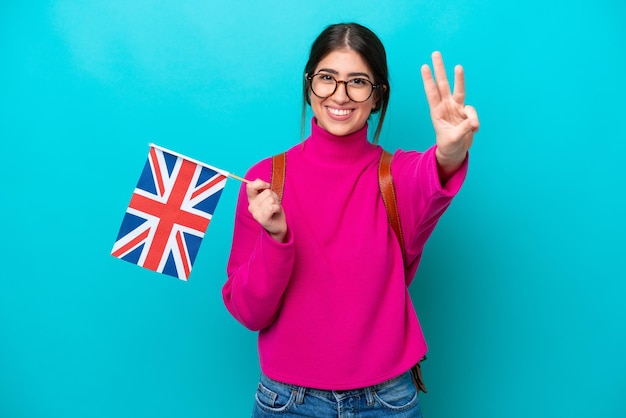 Young caucasian student woman holding English flag isolated on blue background happy and counting three with fingers