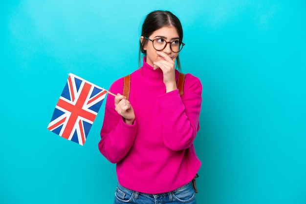 Young caucasian student woman holding English flag isolated on blue background having doubts