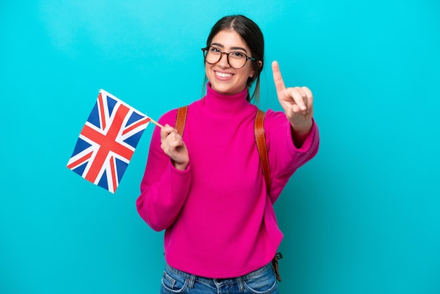 Young caucasian student woman holding English flag isolated on blue background showing and lifting a finger