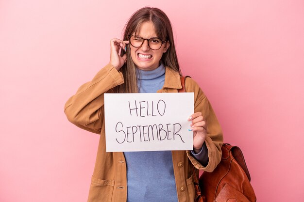 손으로 귀를 덮고 분홍색 배경에 고립 된 안녕하세요 9 월 플래 카드를 들고 젊은 백인 학생 여자.