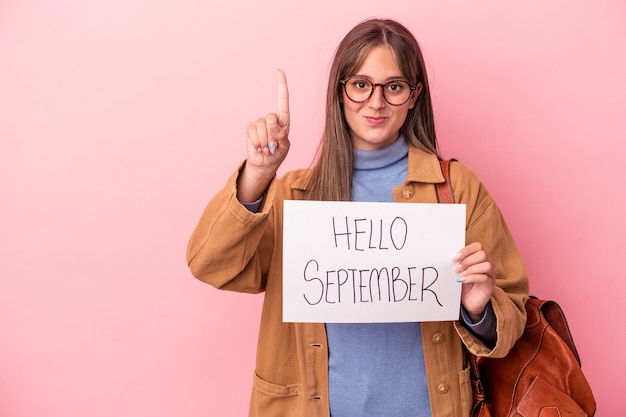 Young caucasian student woman holding hello September placard isolated on pink background showing number one with finger.