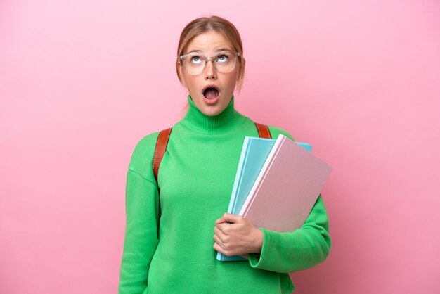 Young caucasian student woman isolated on pink background looking up and with surprised expression