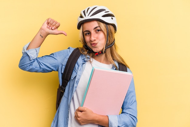 黄色の背景に分離された自転車のヘルメットを身に着けている若い白人学生の女性は、誇りと自信を感じています。