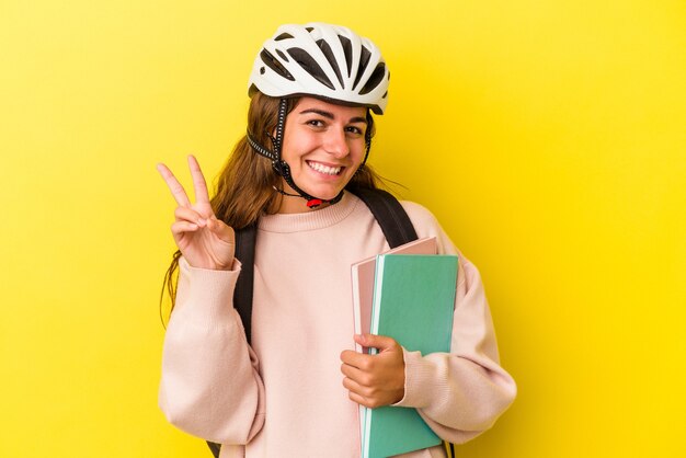 Молодая кавказская студентка женщина в велосипедном шлеме, изолированном на желтом фоне, показывает номер два пальцами.