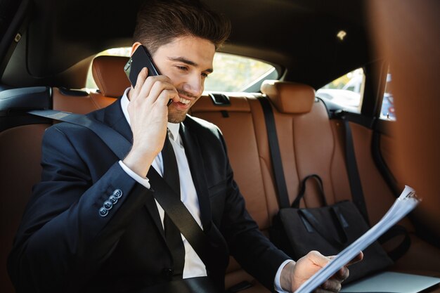 young caucasian successful businesslike man in formal black suit talking on cellphone while riding in car