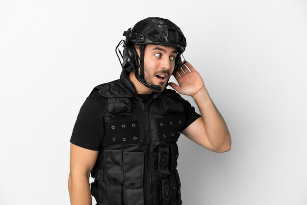 Young caucasian swat isolated on white background listening to something by putting hand on the ear