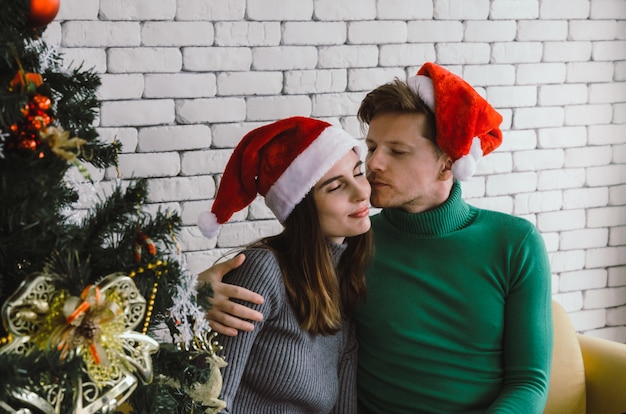 Young caucasian sweet couple with red santa hat enjoy and kissing his girlfriend on sofa with christmas tree celebrating in house