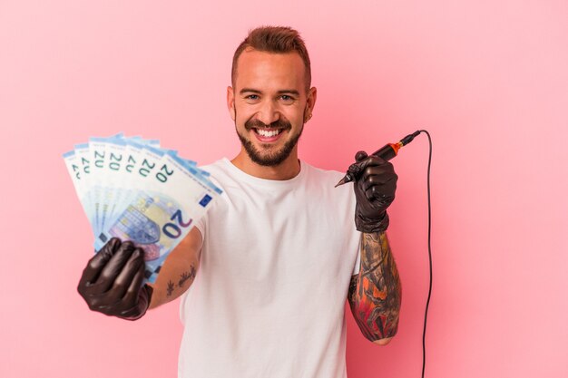Young caucasian tattoo artist holding bills isolated on pink background