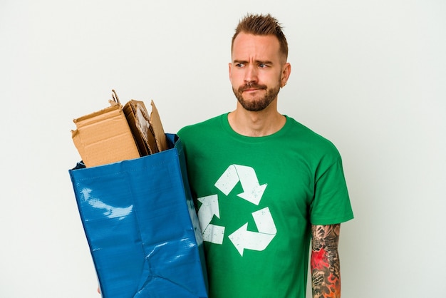 Young caucasian tattooed man recycled cardboard isolated on white background confused, feels doubtful and unsure.