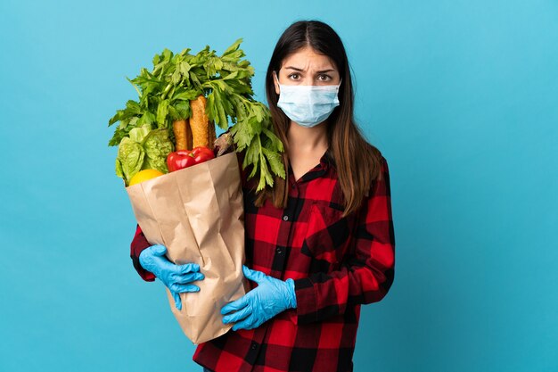 Young caucasian with vegetables and mask isolated on blue with sad expression