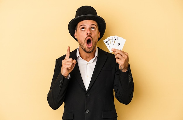 Young caucasian wizard holding magic card isolated on yellow background pointing upside with opened mouth.