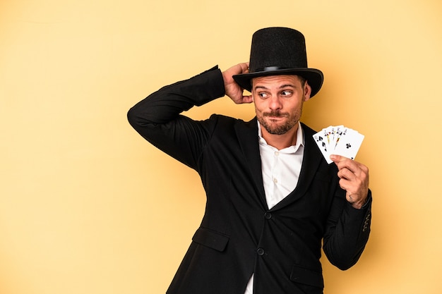 Young caucasian wizard holding magic card isolated on yellow\
background touching back of head, thinking and making a\
choice.