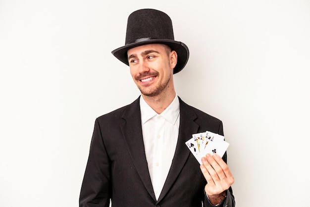 Young caucasian wizard man holding a magic cards isolated on white background looks aside smiling, cheerful and pleasant.