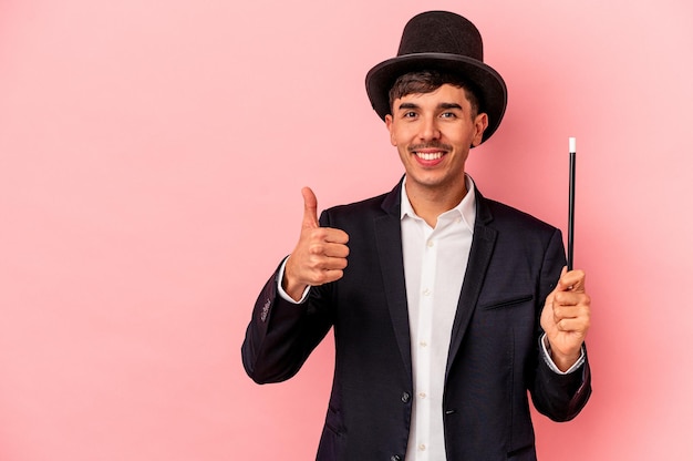 Young caucasian wizard man holding a wand isolated on pink background smiling and raising thumb up