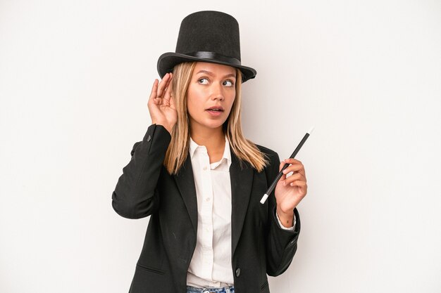 Young caucasian wizard woman holding wand isolated on white background trying to listening a gossip.