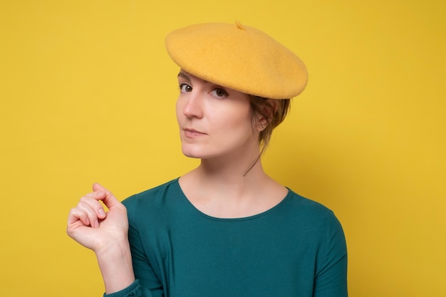 顔に真剣な表情で黄色のベレー帽を身に着けている若い白人のwoamn黄色の壁に撮影されたシンプルで自然に見えるスタジオ