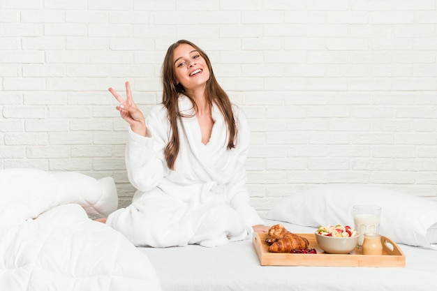 Молодая кавказская женщина на кровати радостная и беззаботная показывая символ мира с пальцами.