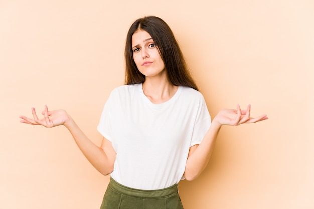 Young caucasian woman on beige wall doubting and shrugging shoulders in questioning gesture.