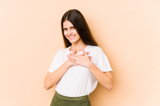 Young caucasian woman on beige wall has friendly expression, pressing palm to chest