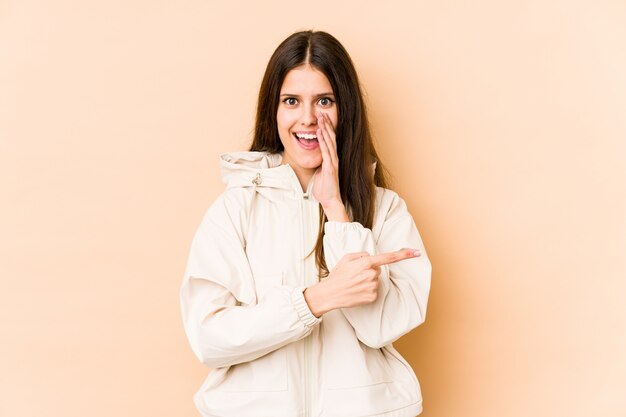 Young caucasian woman on beige wall saying a gossip, pointing to side reporting something.