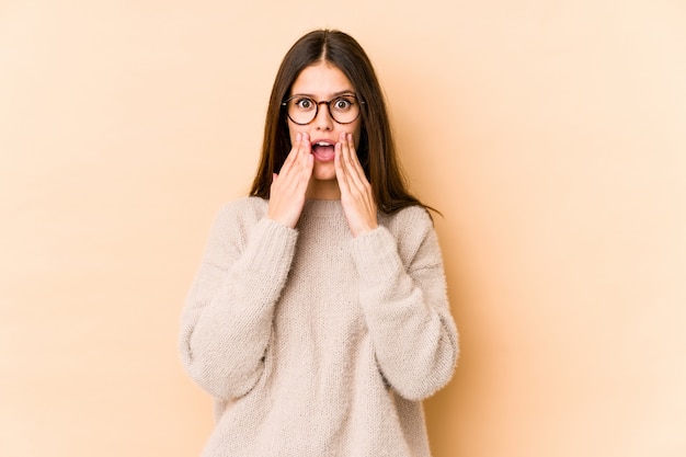 Young caucasian woman on beige wall shocked, covering mouth with hands, anxious to discover something new.