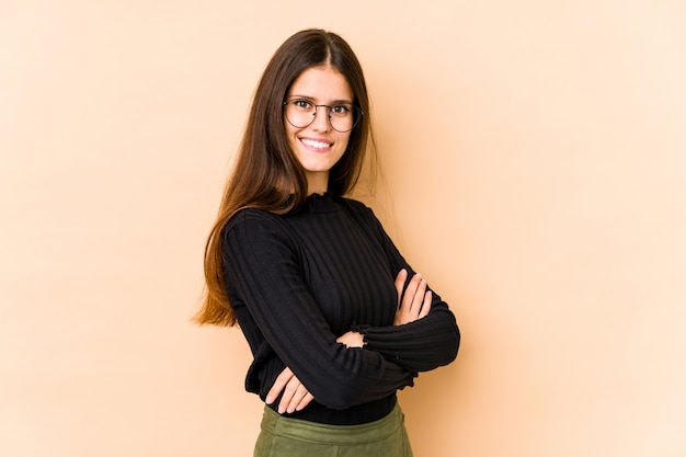 Young caucasian woman on beige wall who feels confident, crossing arms with determination.