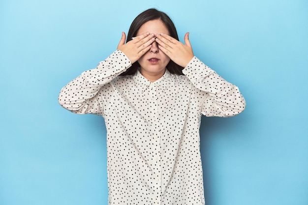 Young Caucasian woman on blue backdrop afraid covering eyes with hands