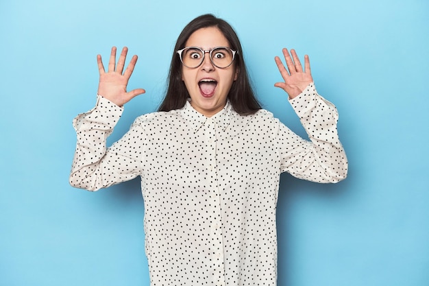 Young caucasian woman on blue backdrop receiving a pleasant surprise excited and raising hands