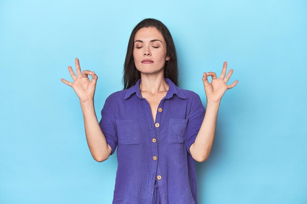 Young caucasian woman on blue backdrop relaxes after hard working day she is performing yoga