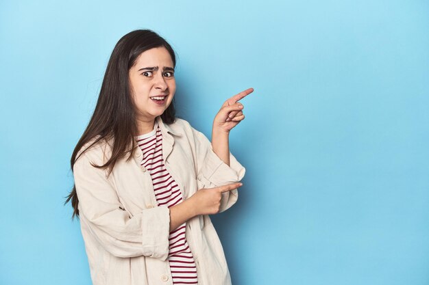 Young caucasian woman on blue backdrop shocked pointing with index fingers to a copy space