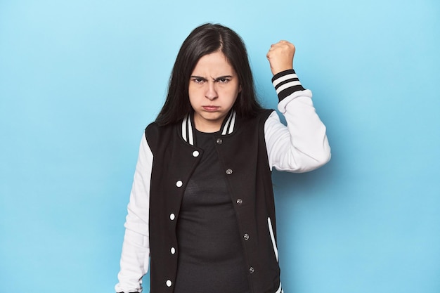 Young caucasian woman on blue backdrop showing fist to camera aggressive facial expression