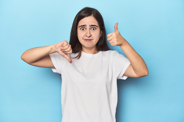 Young Caucasian woman on blue backdrop showing thumbs up and thumbs down difficult choose concept