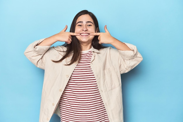 Young Caucasian woman on blue backdrop smiles pointing fingers at mouth