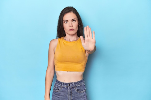 Young caucasian woman on blue backdrop standing with outstretched hand showing stop sign preventing you