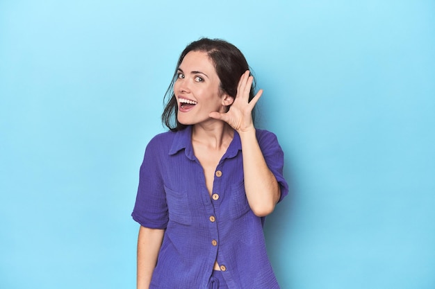 Young caucasian woman on blue backdrop trying to listening a gossip