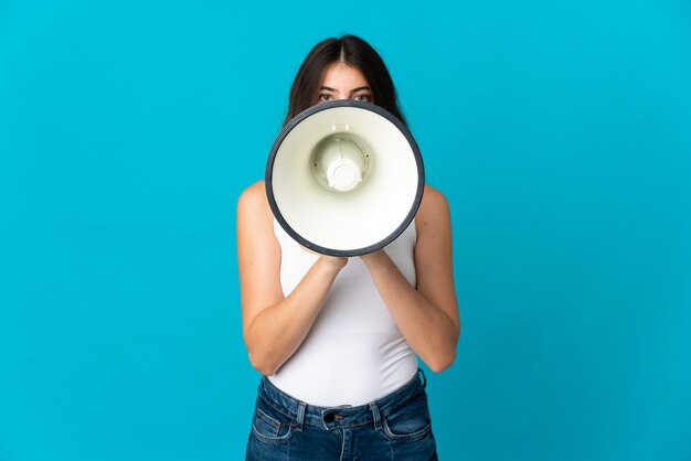 Young caucasian woman on blue shouting through a megaphone to announce something